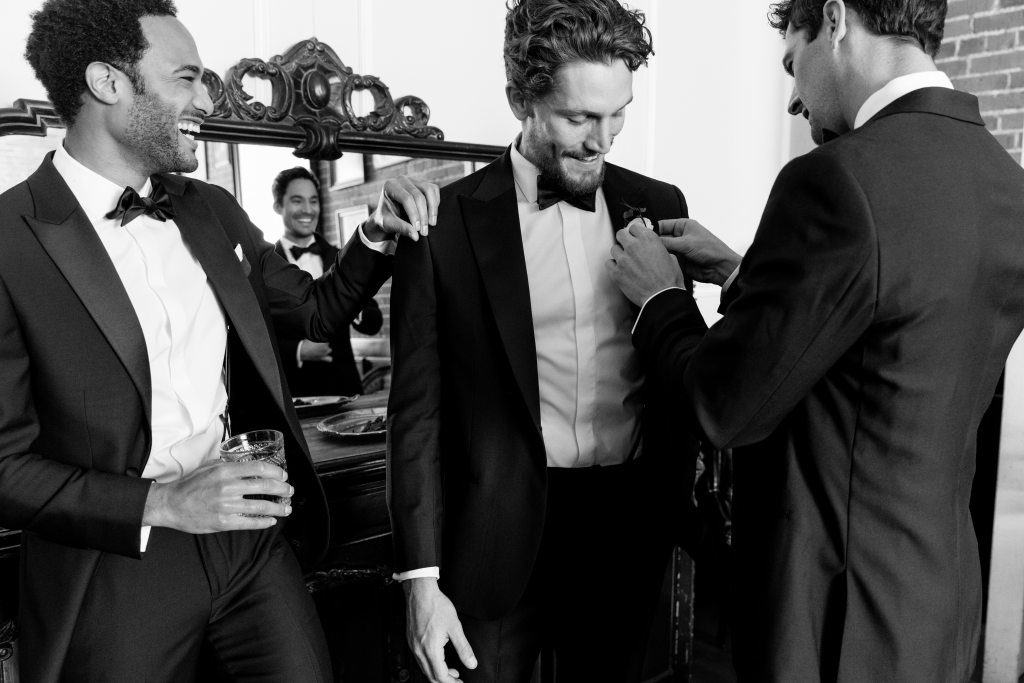 Groomsmen help groom prepare in black tie optional tuxedos by The Black Tux.