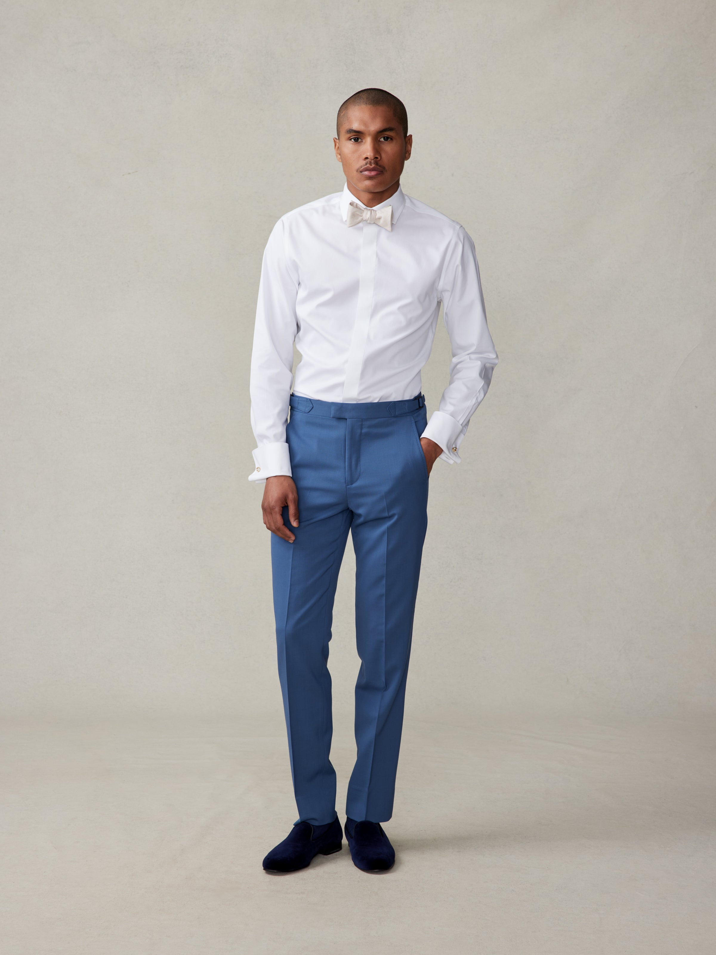 Man wearing a white shirt, blue trousers, and a bow tie, standing against a neutral background.