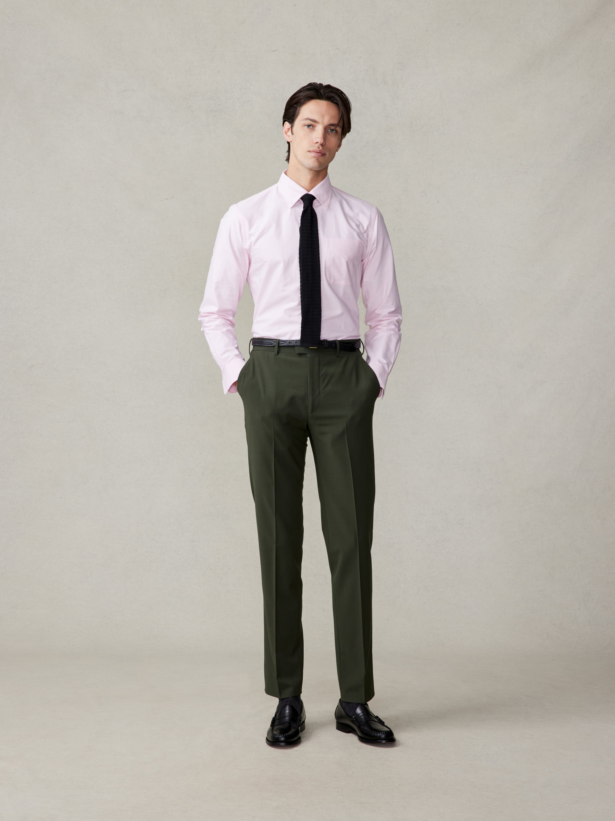 Man wearing a pink shirt, black tie, and green trousers, standing on a neutral background.