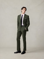 A man wearing a tailored olive green suit, a light pink oxford, and a black tie, standing confidently with his hands in his pockets.