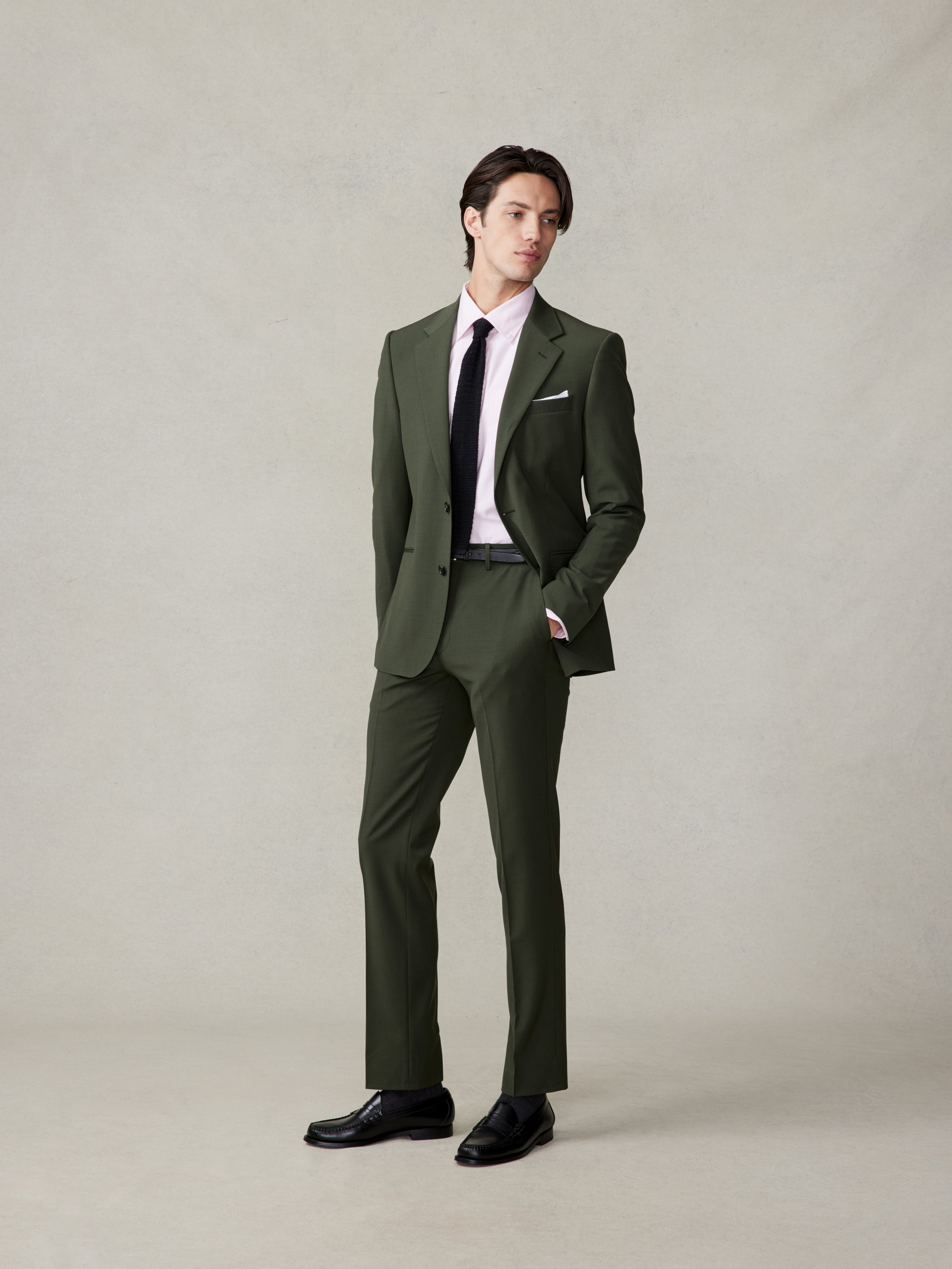 A man wearing a tailored olive green suit, a light pink oxford, and a black tie, standing confidently with his hands in his pockets.