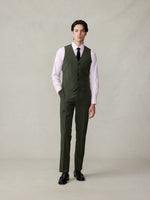 A man wearing an olive green suit vest over a pink oxford shirt and black tie, standing against a neutral background.