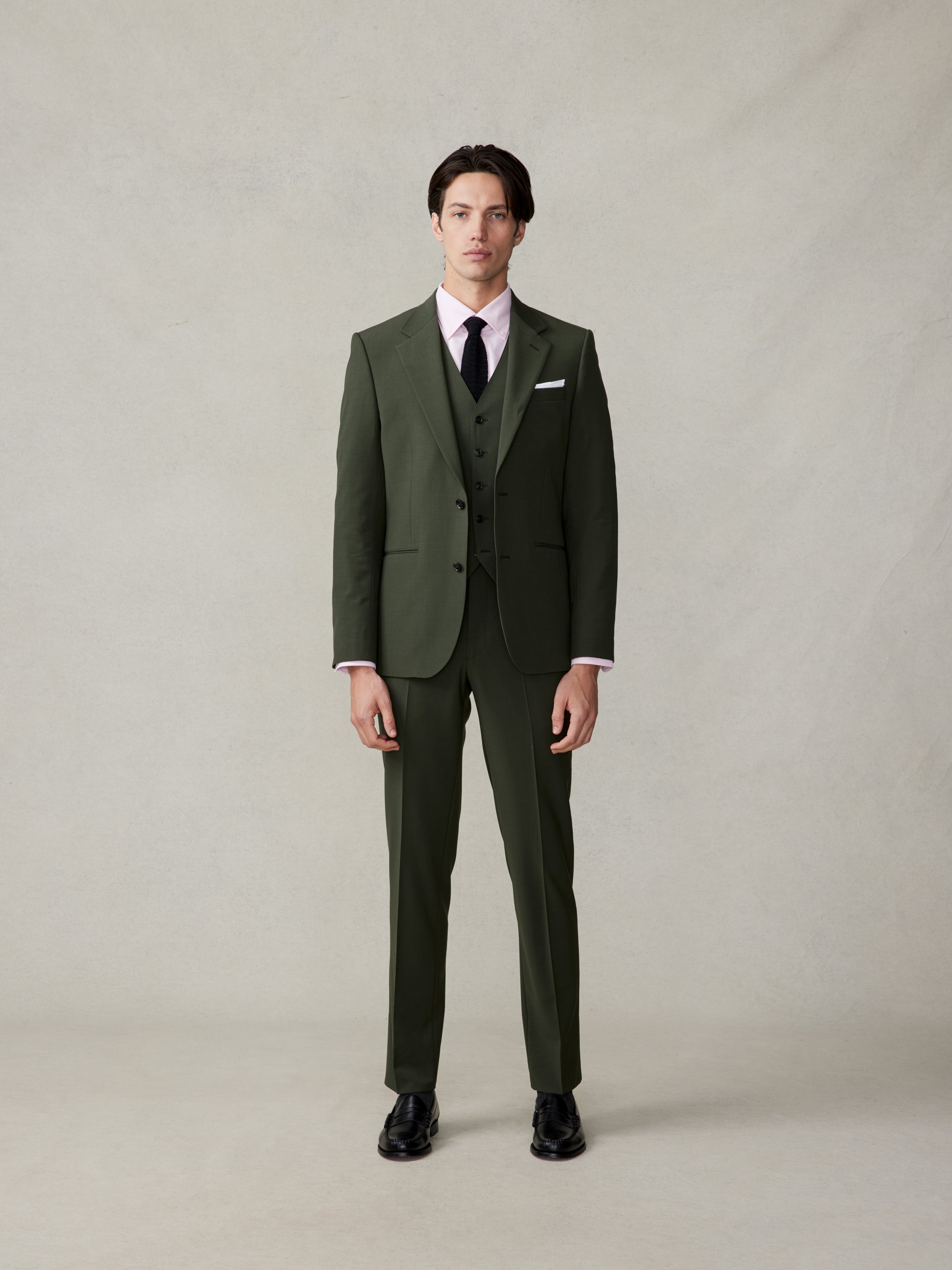 Man wearing a green suit with a pink shirt and black tie, standing against a neutral background.