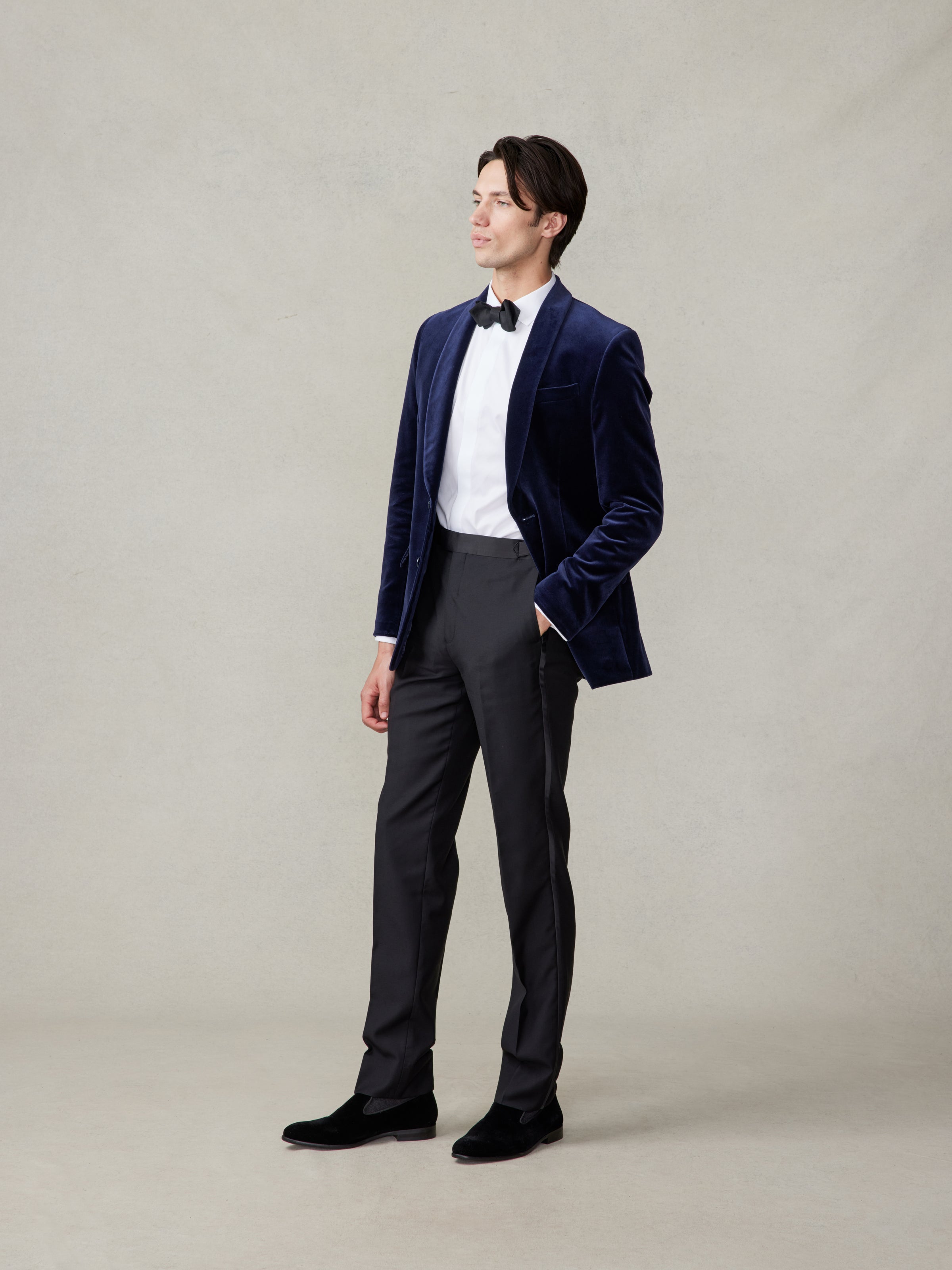 Man in ocean blue velvet tuxedo jacket with black tuxedo pants, side profile, looking ahead, on beige backdrop
