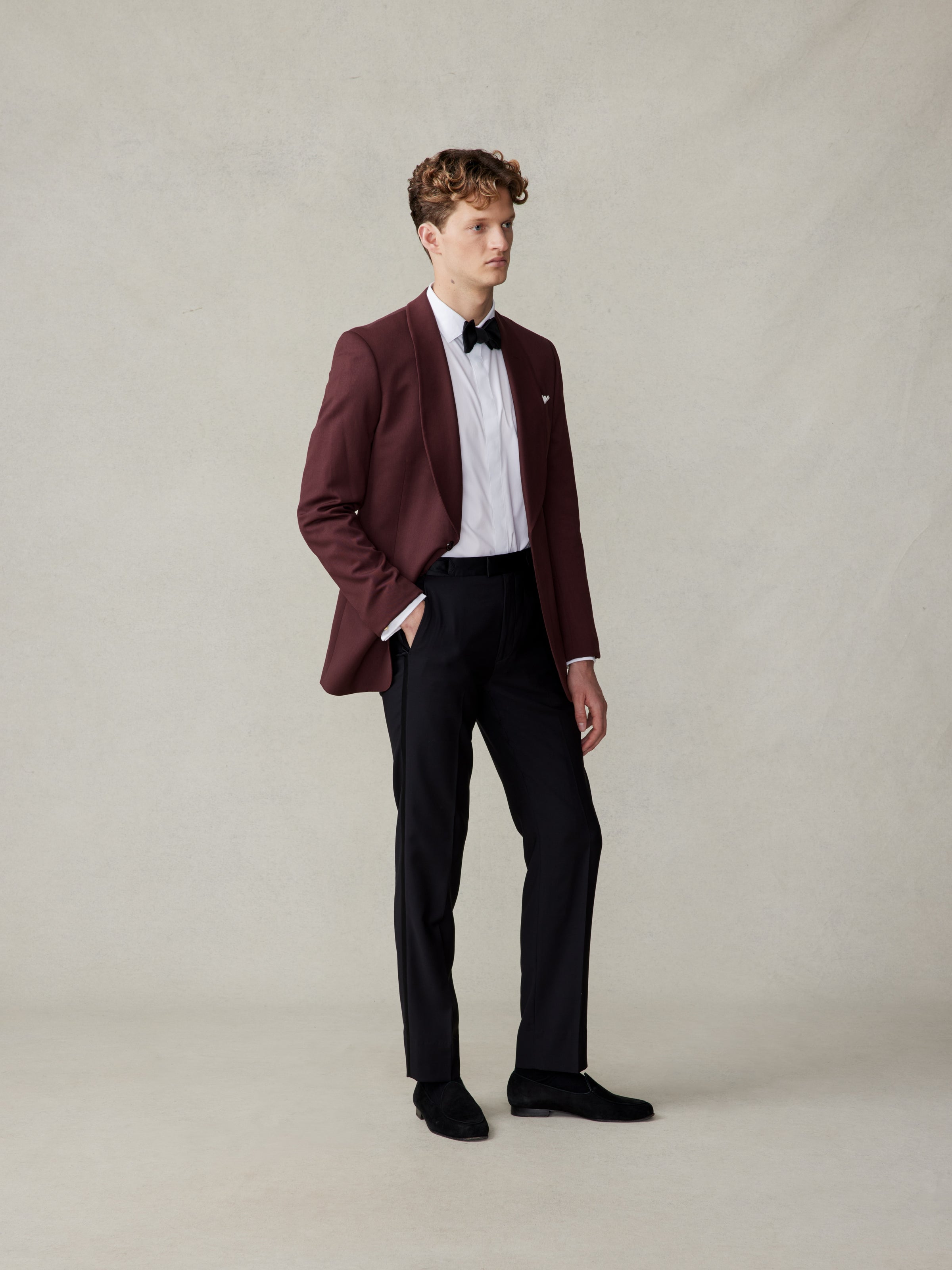 Man in a burgundy tuxedo jacket, white shirt, and black pants standing against a neutral background.