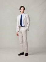 Man in a light beige suit with a blue shirt and tie, standing against a neutral background.