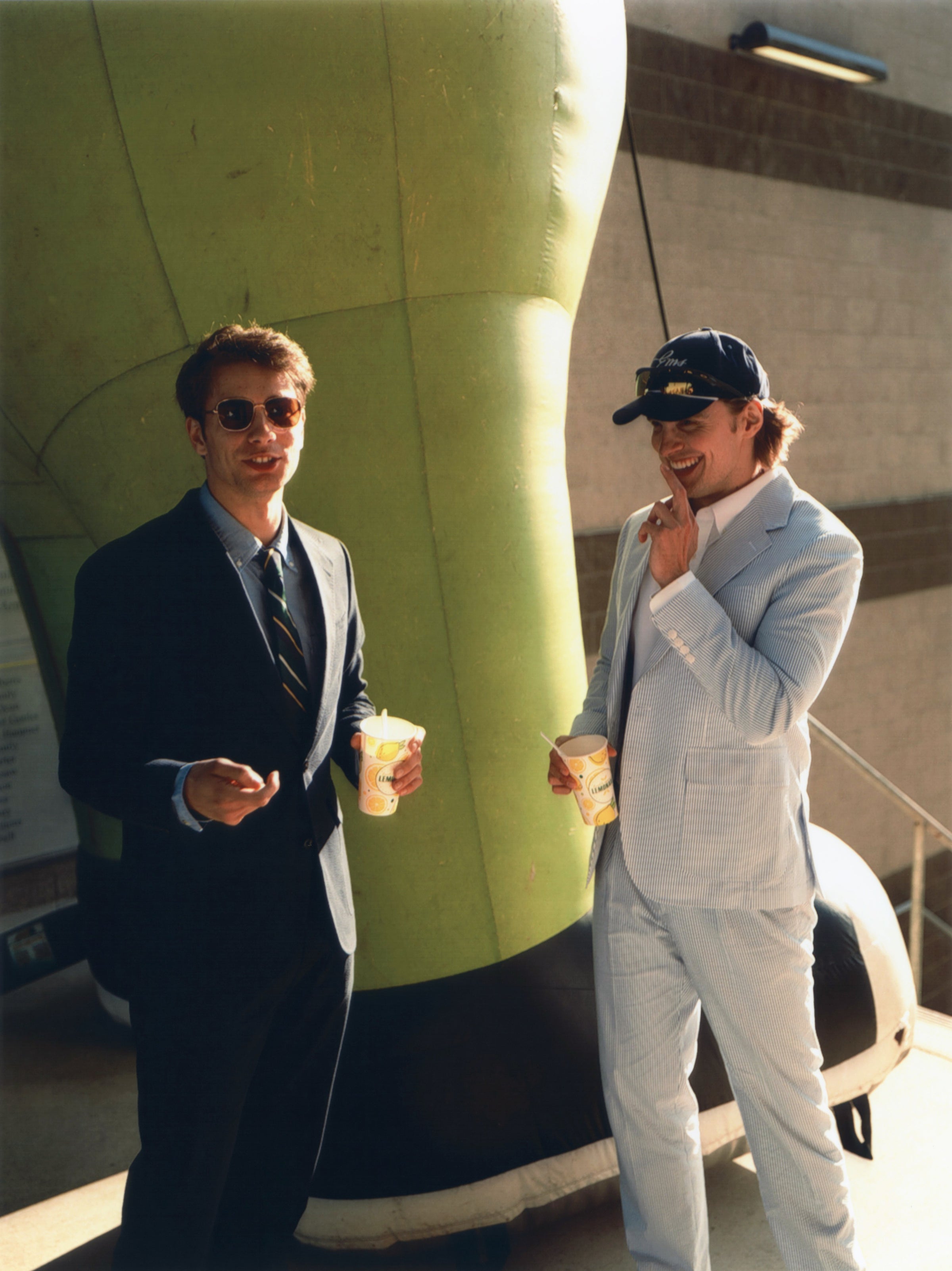 Two men in suits holding drinks, standing in front of a large green inflatable object.