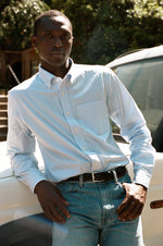Man in a striped blue shirt and jeans leaning against a white vehicle.