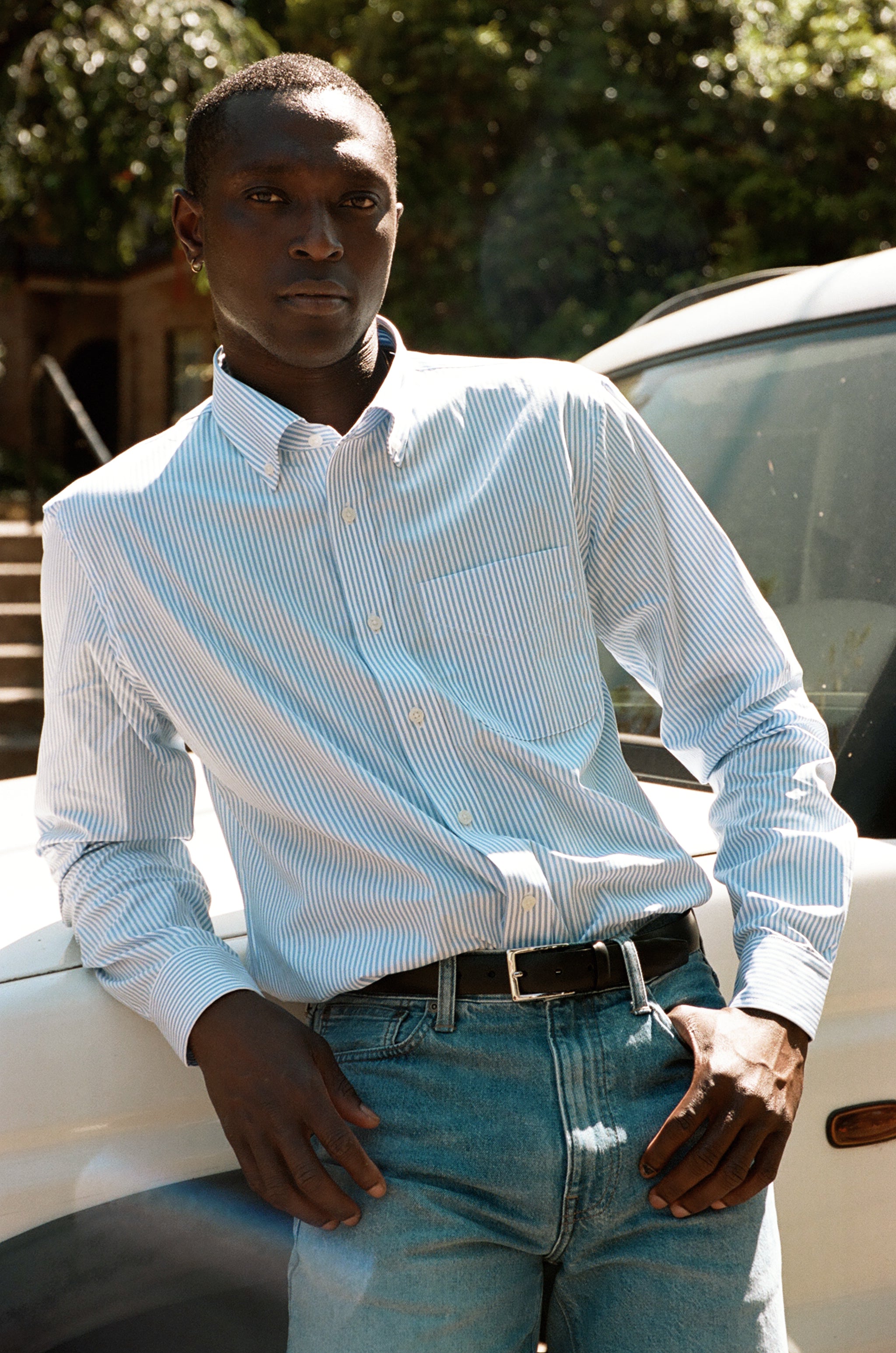 Man in a striped blue shirt and jeans leaning against a white vehicle.