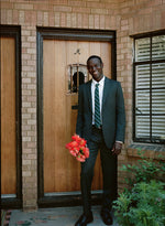 Man in a dark suit holding a bouquet of red flowers, standing by a wooden door.