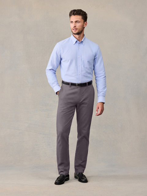 Man in a light blue button-down shirt and grey chino pants, standing against a tan backdrop.