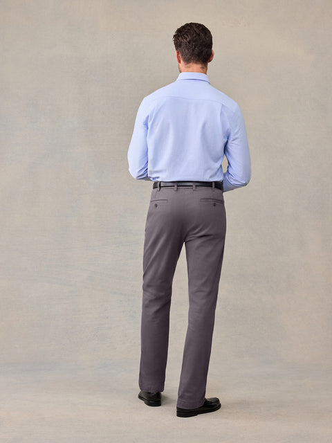 Back view of a man in a light blue shirt and grey chino pants against a tan backdrop.