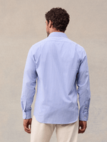 Back view of a man in a light blue and white wide stripe dress shirt with cream trousers, beige backdrop.