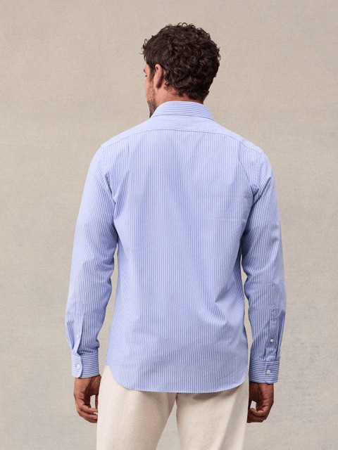 Back view of a man in a light blue and white wide stripe dress shirt with cream trousers, beige backdrop.