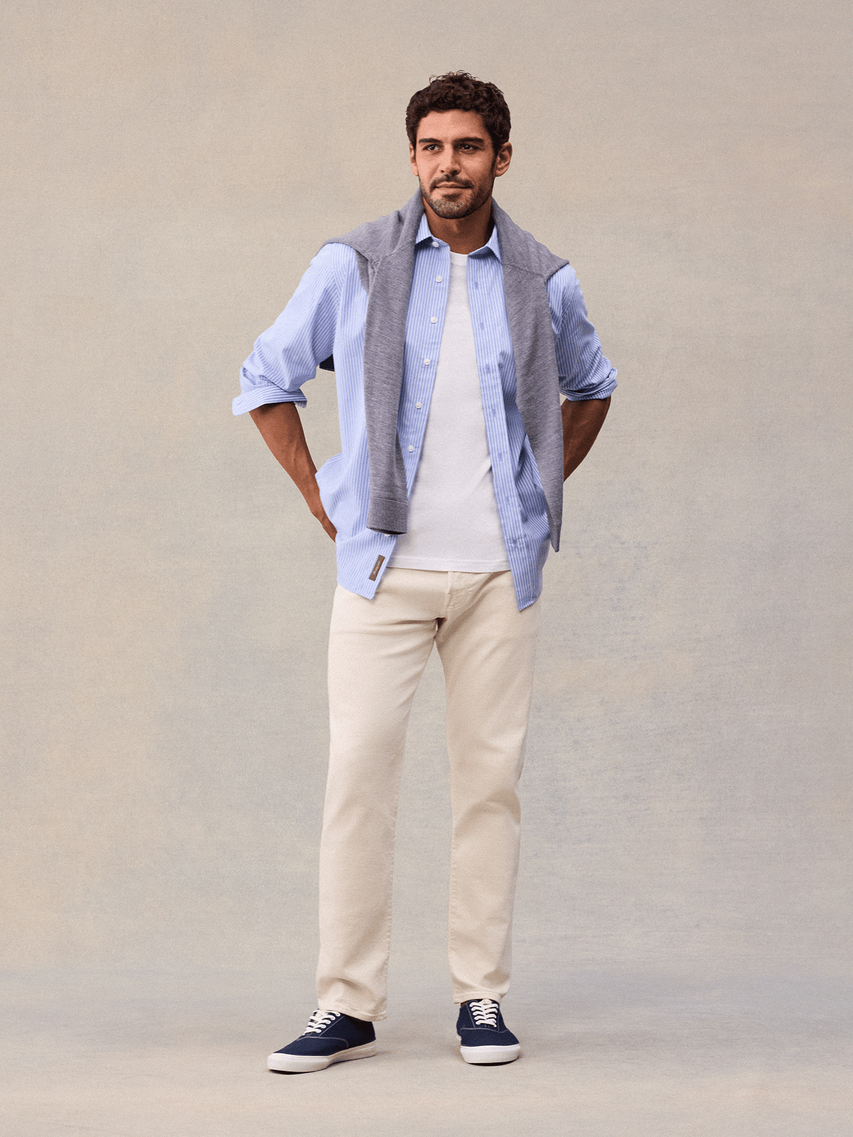 A man in a light blue and white wide stripe dress shirt, sleeves rolled, grey sweater on shoulders, navy sneakers.