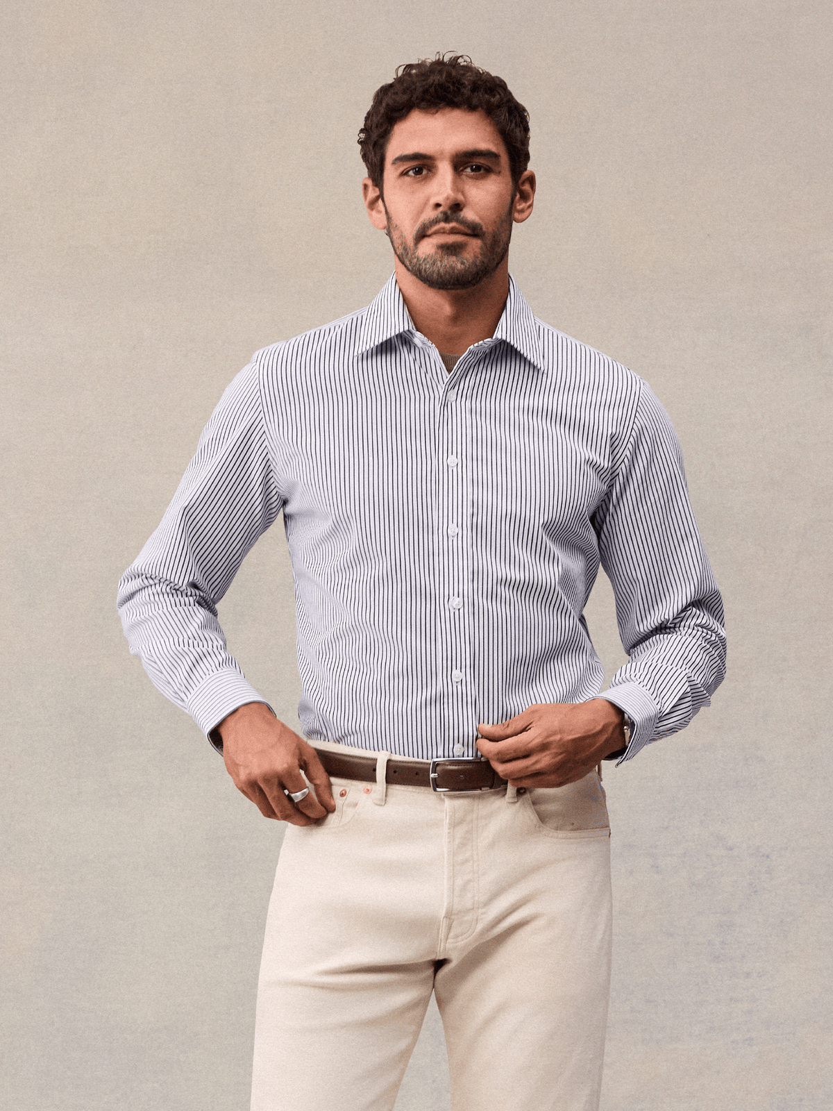 A man in a white and navy wide stripe dress shirt tucked into cream trousers with a brown belt, front view.