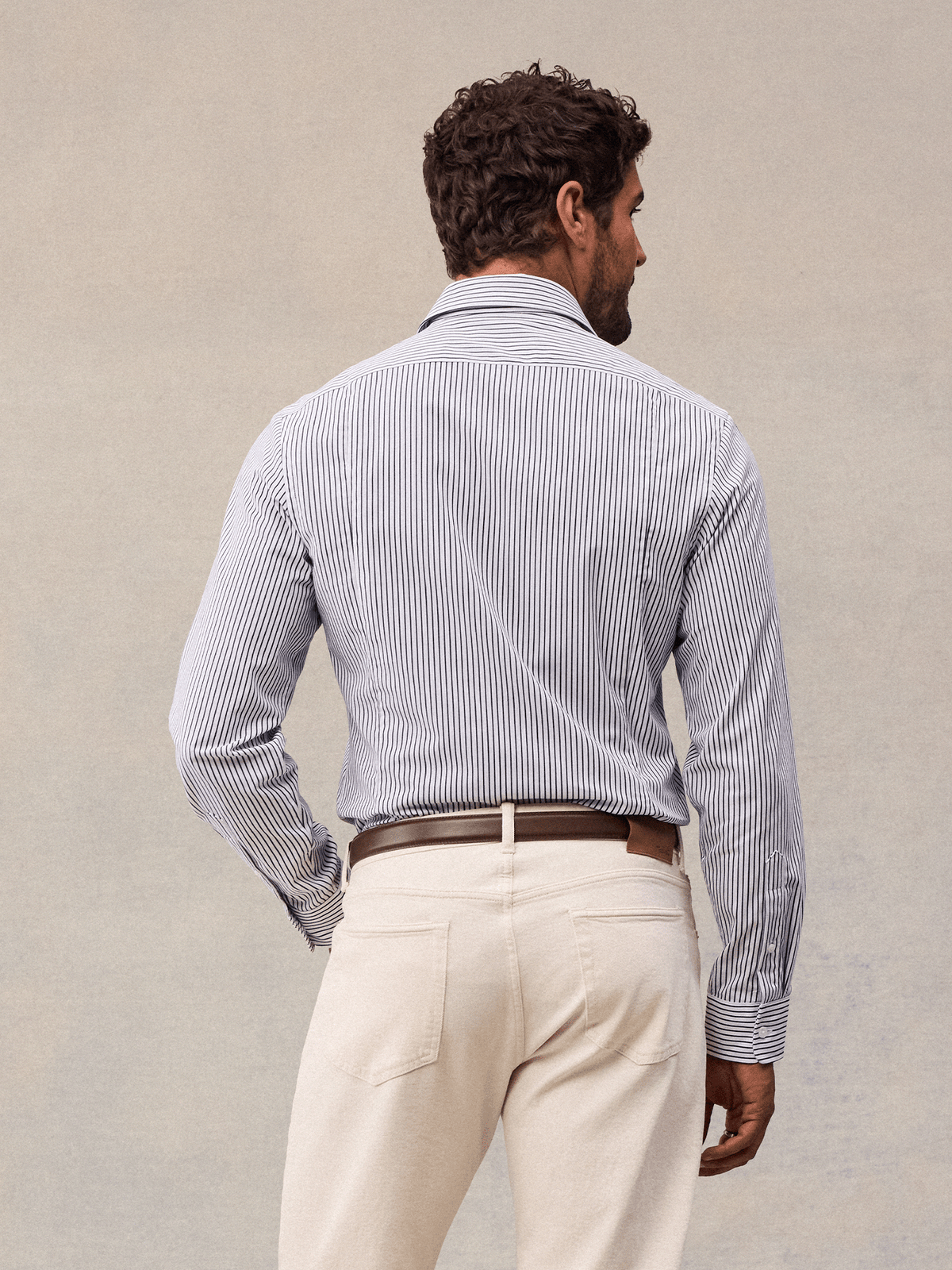 Back view of a man in a white and navy wide stripe dress shirt, cream trousers and brown belt, beige backdrop.