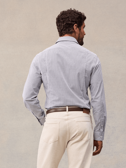 Back view of a man in a white and navy wide stripe dress shirt, cream trousers and brown belt, beige backdrop.