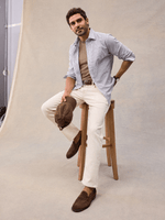 A man seated on a stool in an open white and navy wide stripe dress shirt over a tan tee, cream trousers, suede loafers.