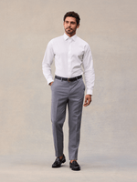 A man in slate grey suit trousers, white dress shirt and black belt, full length, black tassel loafers.