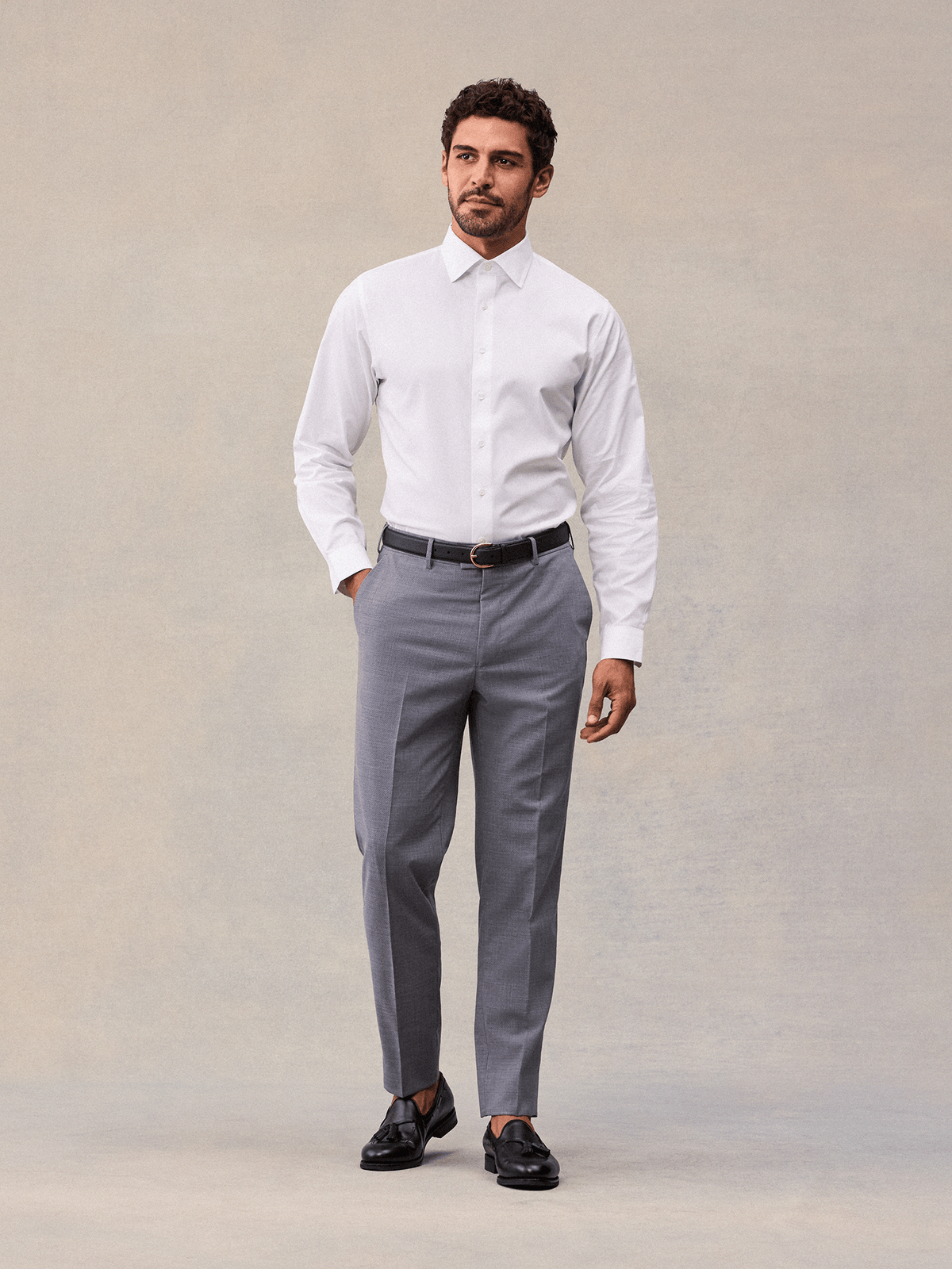 A man in slate grey suit trousers, white dress shirt and black belt, full length, black tassel loafers.