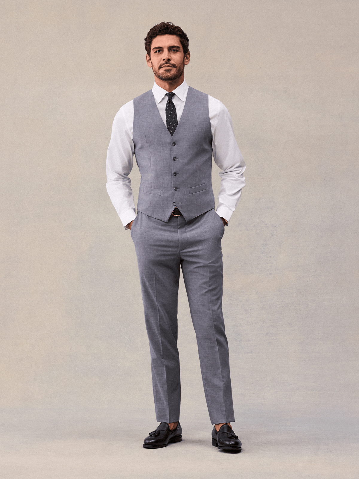 A man in a slate grey suit vest and matching trousers, full length, white shirt and black tassel loafers.