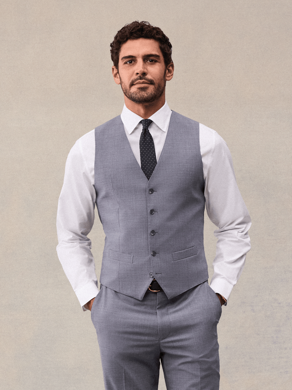 A man in a slate grey suit vest with welt pockets, white shirt and navy pindot tie, beige backdrop.