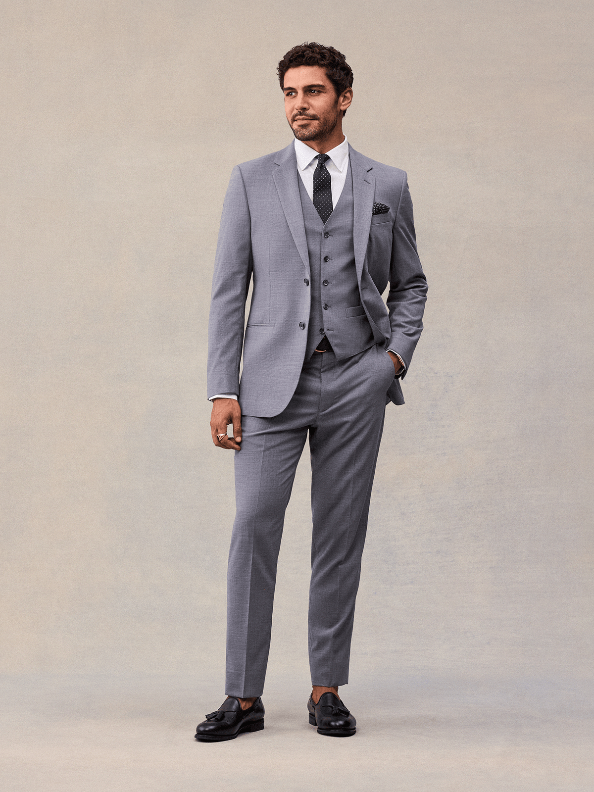 A man in a slate grey three-piece suit with navy pindot tie and pocket square, full length, looking away.