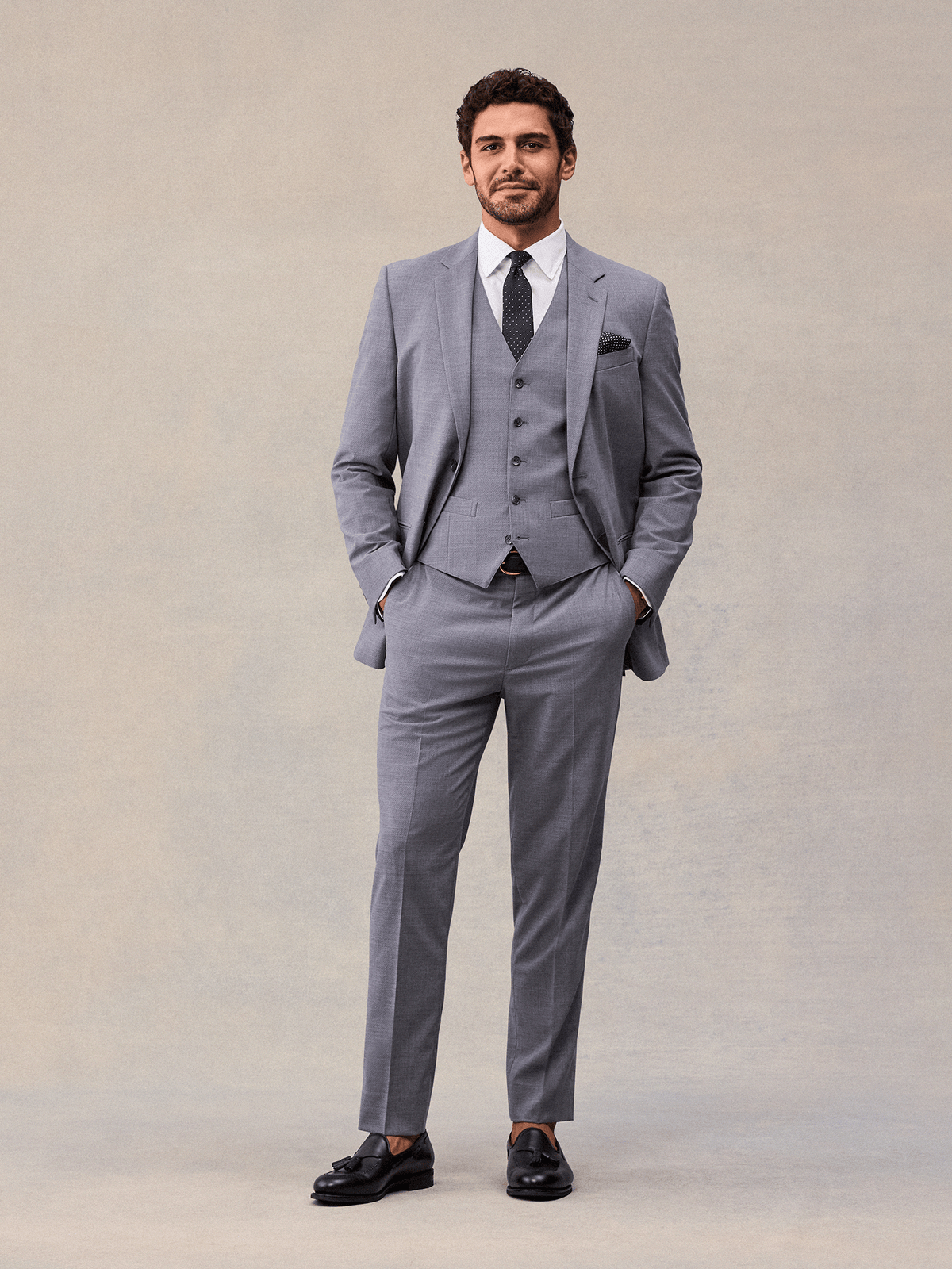 A man in a slate grey three-piece suit, jacket open over the vest, hands in pockets, black tassel loafers.