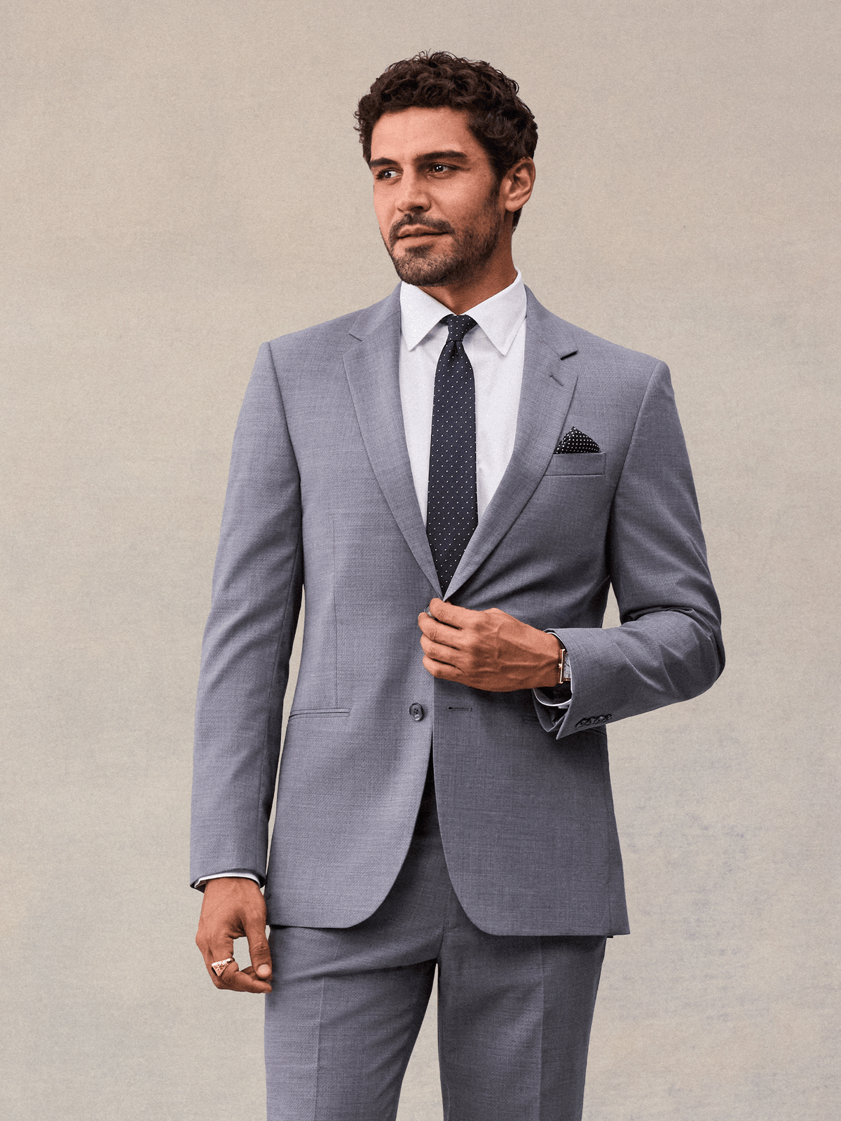 A man buttoning a slate grey suit jacket with notch lapels, white shirt and navy pindot tie, beige backdrop.