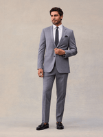 A man in a slate grey suit buttoning the jacket, full length, navy pindot tie and black tassel loafers.