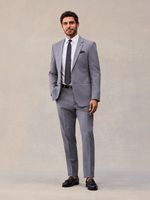 A man in a slate grey two-piece suit, full length, hand in pocket, navy pindot tie and black tassel loafers.