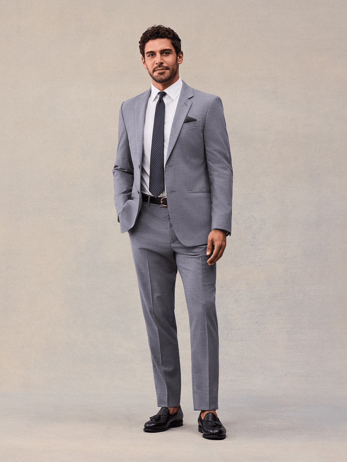 A man in a slate grey two-piece suit, full length, hand in pocket, navy pindot tie and black tassel loafers.