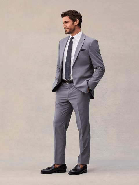 A man in a slate grey suit, three-quarter view, hands in pockets, navy pindot tie and black tassel loafers.