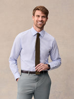 A man in a blue windowpane check dress shirt with an olive knit tie, brown belt and light grey trousers, beige backdrop.