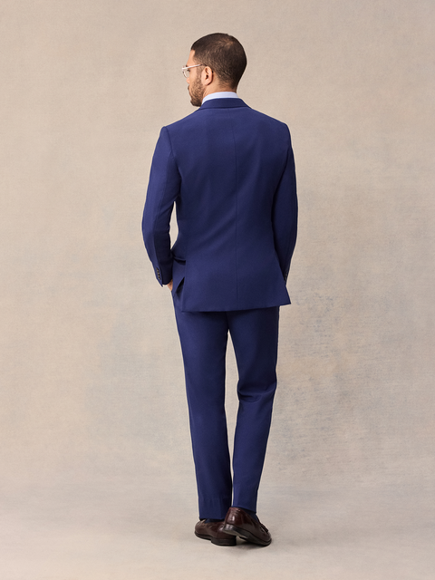 Back view of a man in a cove blue suit jacket and matching suit pants, full length, beige backdrop.
