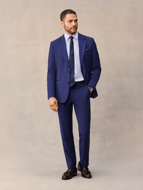 A man in a cove blue suit with the jacket worn open, navy tie, brown tassel loafers, full length.