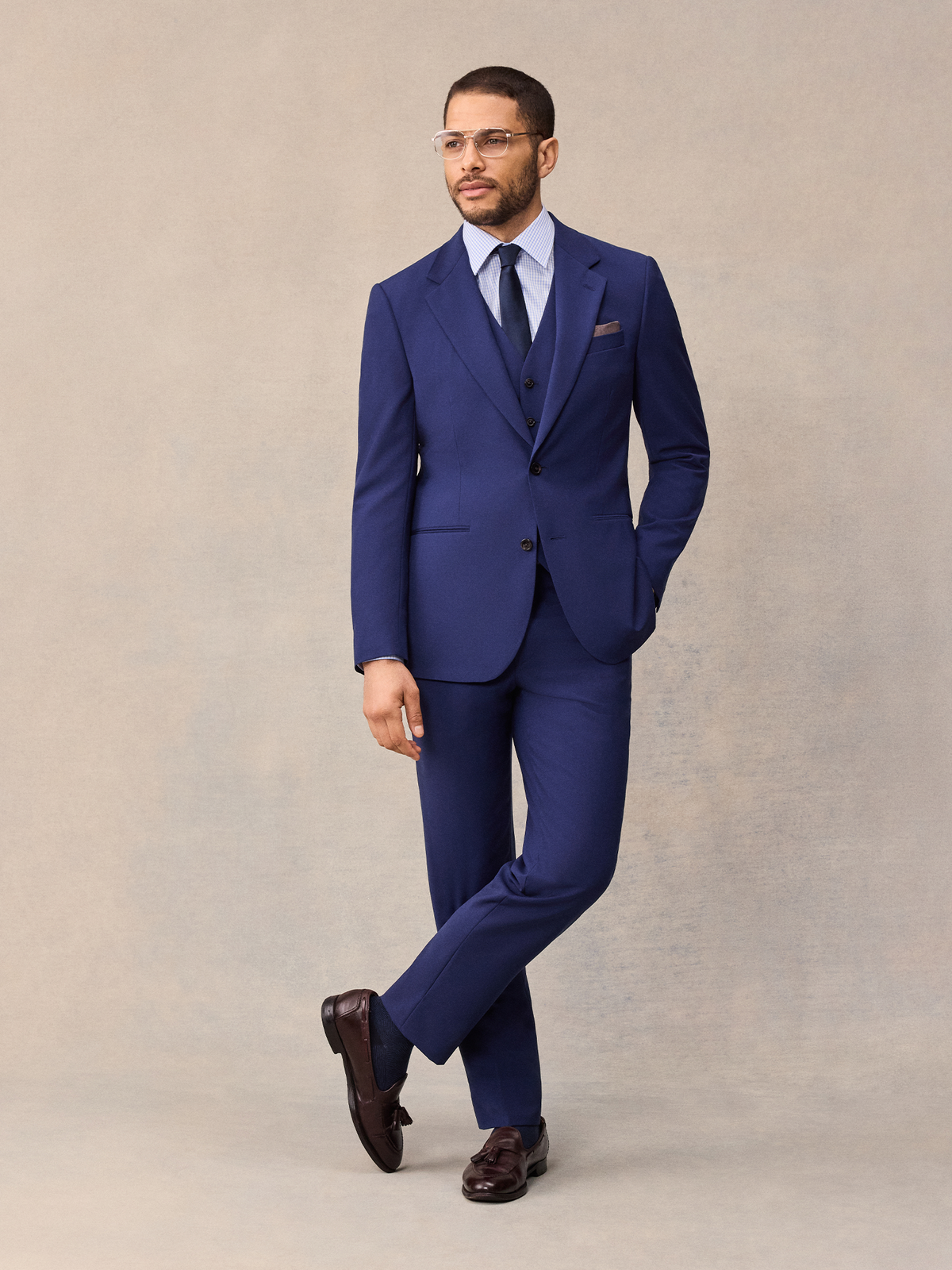 A man in a three-piece cove blue suit with matching vest, navy tie and glasses, legs crossed, full length.