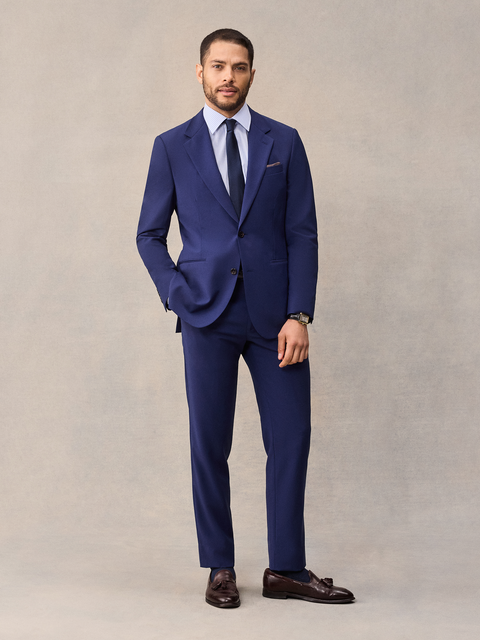 A man in a cove blue suit, full length, hand in pocket, navy tie and brown tassel loafers, beige backdrop.