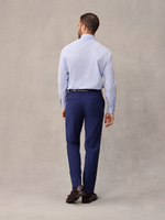Back view of a man in cove blue suit pants and a light blue dress shirt, full length, beige backdrop.