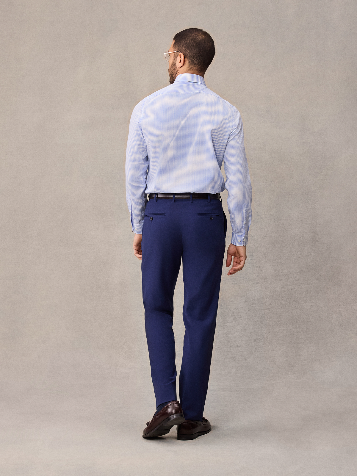 Back view of a man in cove blue suit pants and a light blue dress shirt, full length, beige backdrop.