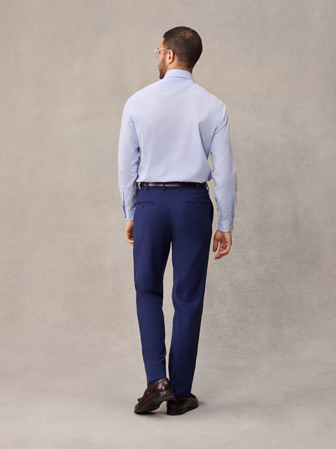 Back view of a man in cove blue suit pants and a light blue dress shirt, full length, beige backdrop.