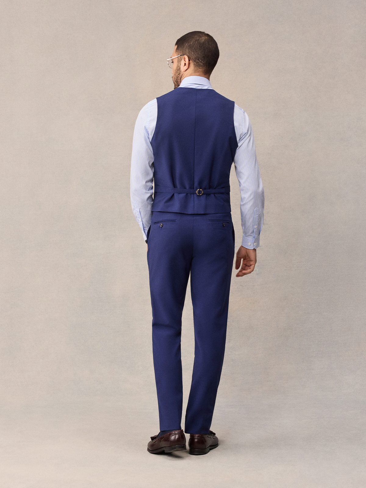 Back view of a man in a cove blue suit vest and matching suit pants, full length, beige backdrop.