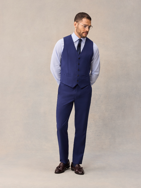 A man in a cove blue suit vest and matching suit pants, navy tie, hands behind back, full length.