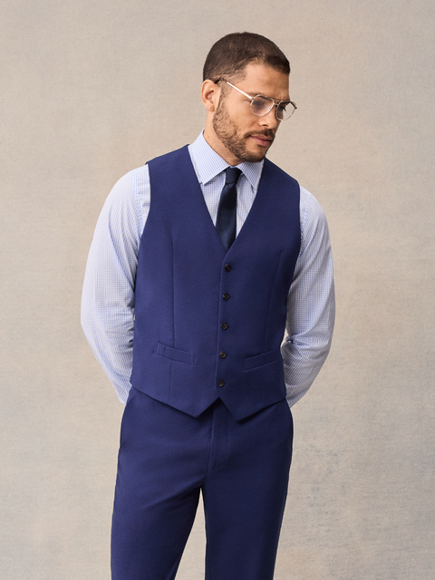 A man in a cove blue suit vest over a light blue shirt with a navy tie and glasses, cropped front.