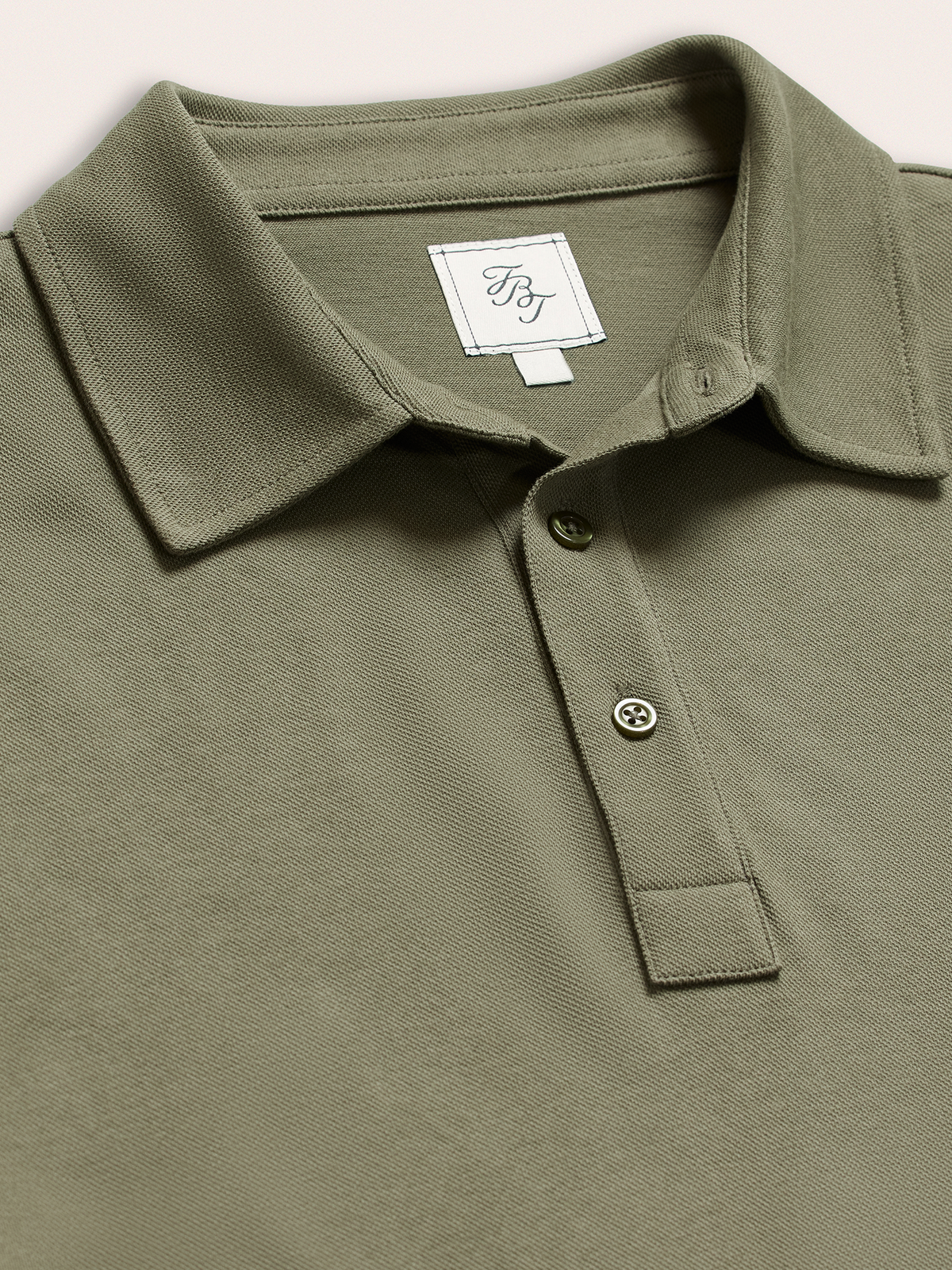 Close-up of olive green pique polo collar and buttons with brand tag, on cream background