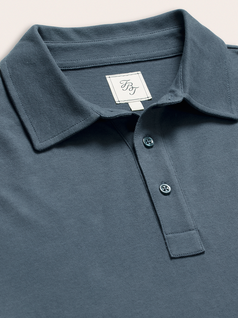 Close-up of slate blue pique polo collar and buttons with brand tag, on cream background