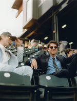Two men sitting in stadium seats, one in a light suit and the other in a dark suit with sunglasses.