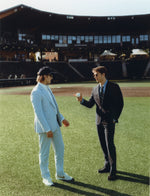 Two men on a baseball field, one in a light suit and the other in a dark suit, holding a baseball.