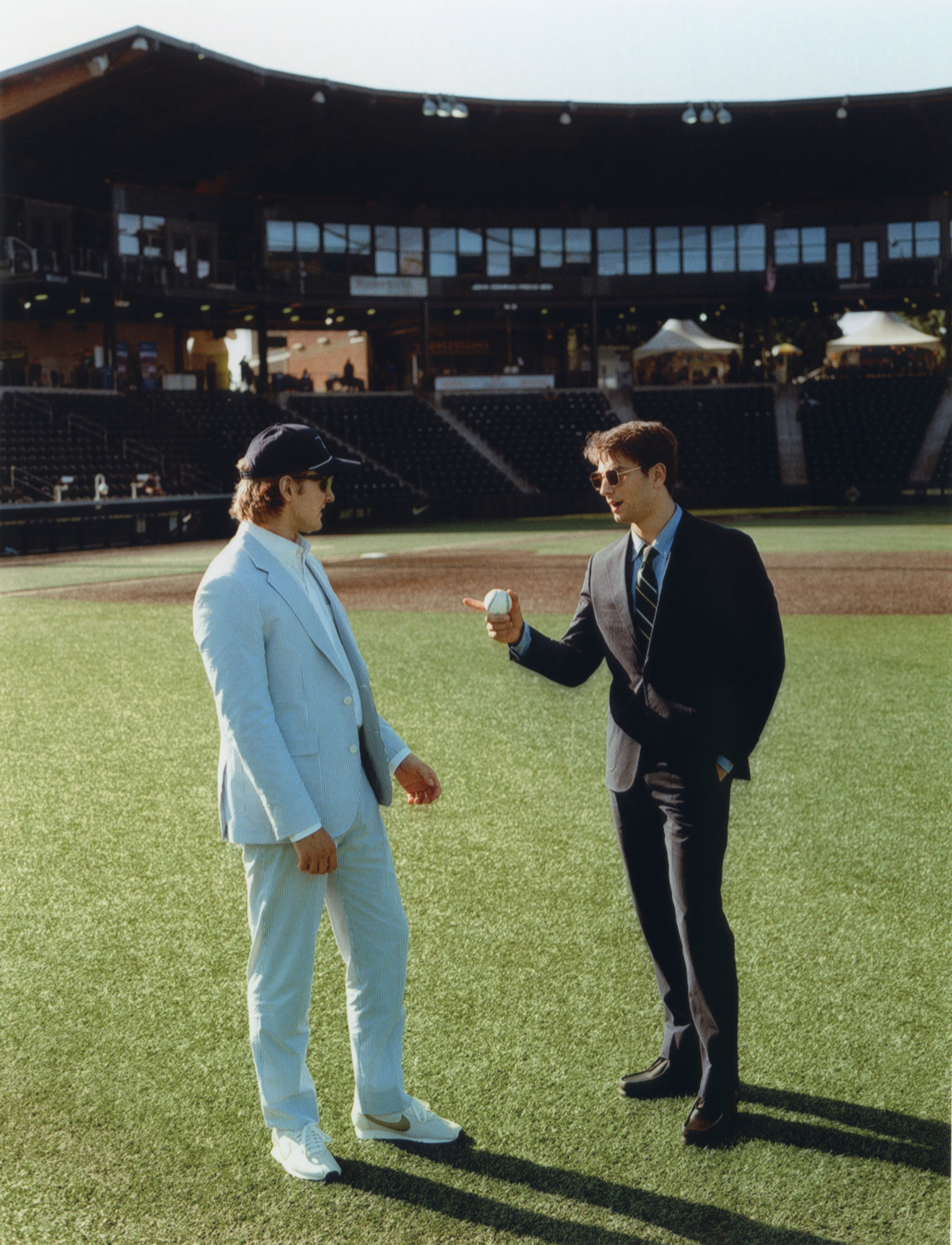 Two men on a baseball field, one in a light suit and the other in a dark suit, holding a baseball.